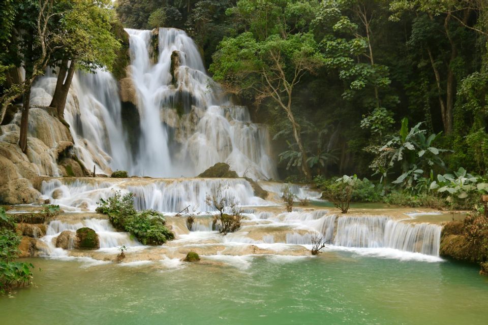 Una cascata a più livelli si getta su rocce chiare in una piscina naturale turchese, avvolta da una lussureggiante foresta verde.