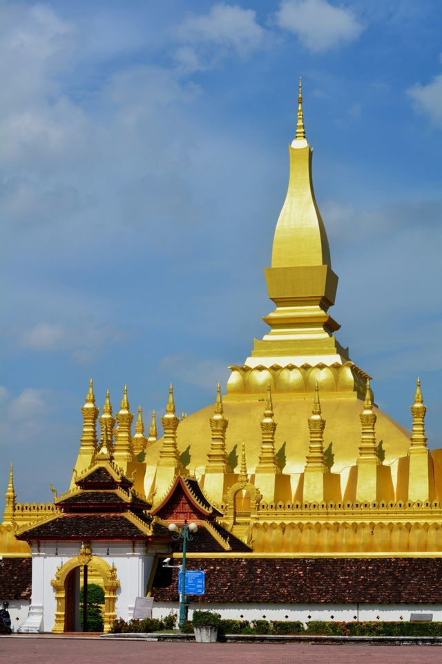 Un grande stupa ricoperto d'oro con molteplici guglie si erge dietro un cancello tradizionale sotto un cielo azzurro con nuvole leggere.