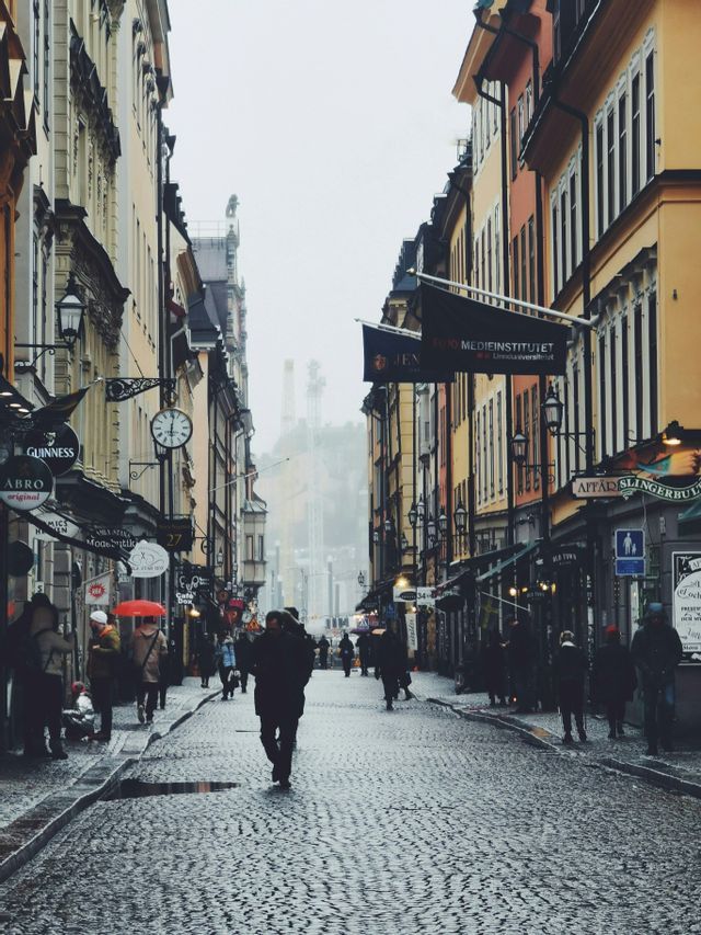 La gente cammina lungo una stretta via acciottolata e bagnata, fiancheggiata da edifici multicolori in una giornata nebbiosa e coperta.