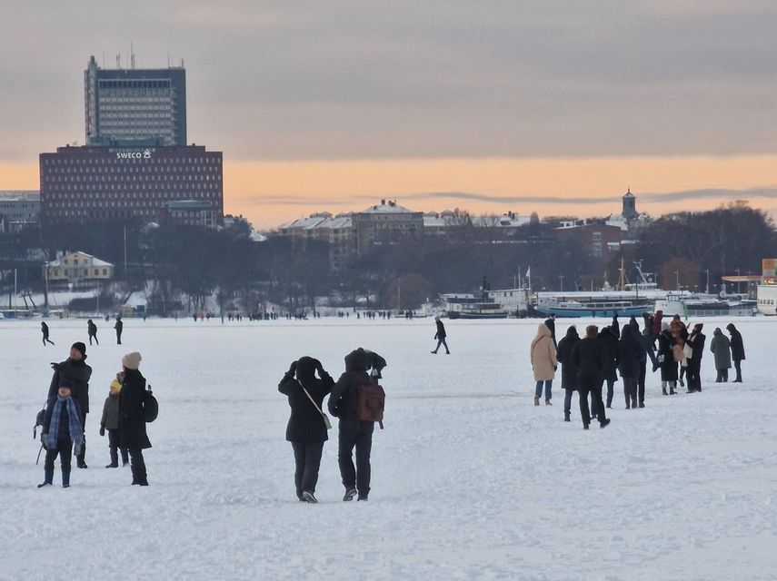 Gruppi di persone con cappotti invernali camminano attraverso una vasta distesa ghiacciata e innevata con lo skyline di una città sullo sfondo al crepuscolo.