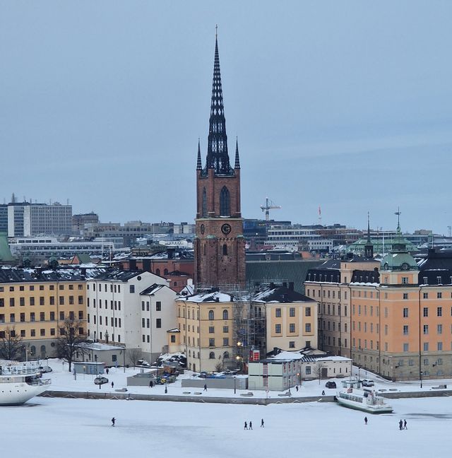Un campanile storico si affaccia su una città coperta di neve e un fiume ghiacciato dove le persone camminano.