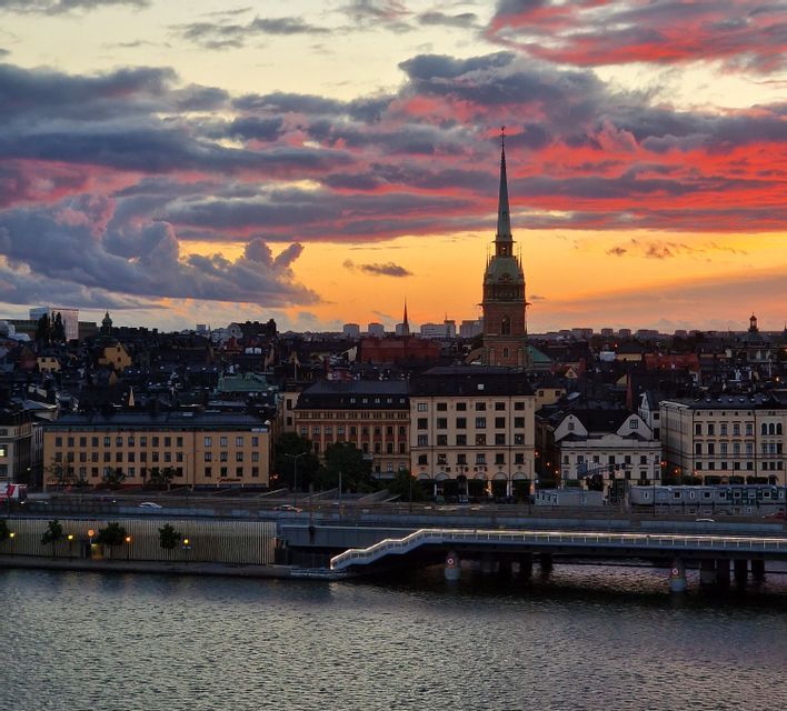Un paesaggio urbano con un prominente campanile di chiesa si erge sulla riva di un fiume, sotto un cielo al tramonto dai colori vivaci, con nuvole arancioni e viola.
