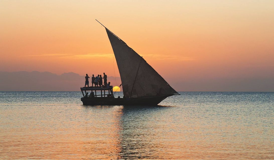 Un gruppo WeRoad in controluce sul ponte di una barca a vela, mentre ammira il vibrante tramonto arancione sull'oceano calmo.