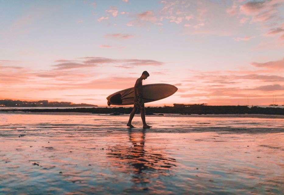 Un surfista cammina con la sua tavola su una spiaggia bagnata, mentre il cielo colorato del tramonto si riflette sulla sabbia.