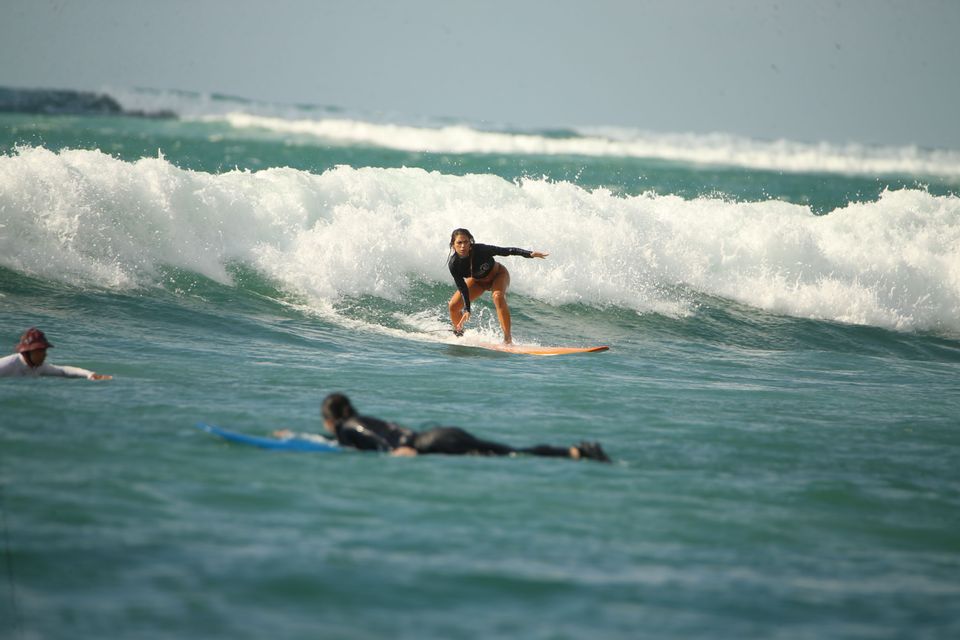 Una donna con una muta nera cavalca un'onda sulla sua tavola da surf, con altri due surfisti in acqua nelle vicinanze.
