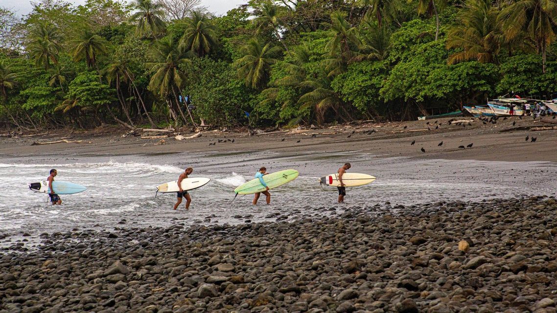Quattro surfisti di un gruppo WeRoad, con le loro tavole, escono dall'acqua su una spiaggia rocciosa con una fitta giungla sullo sfondo.