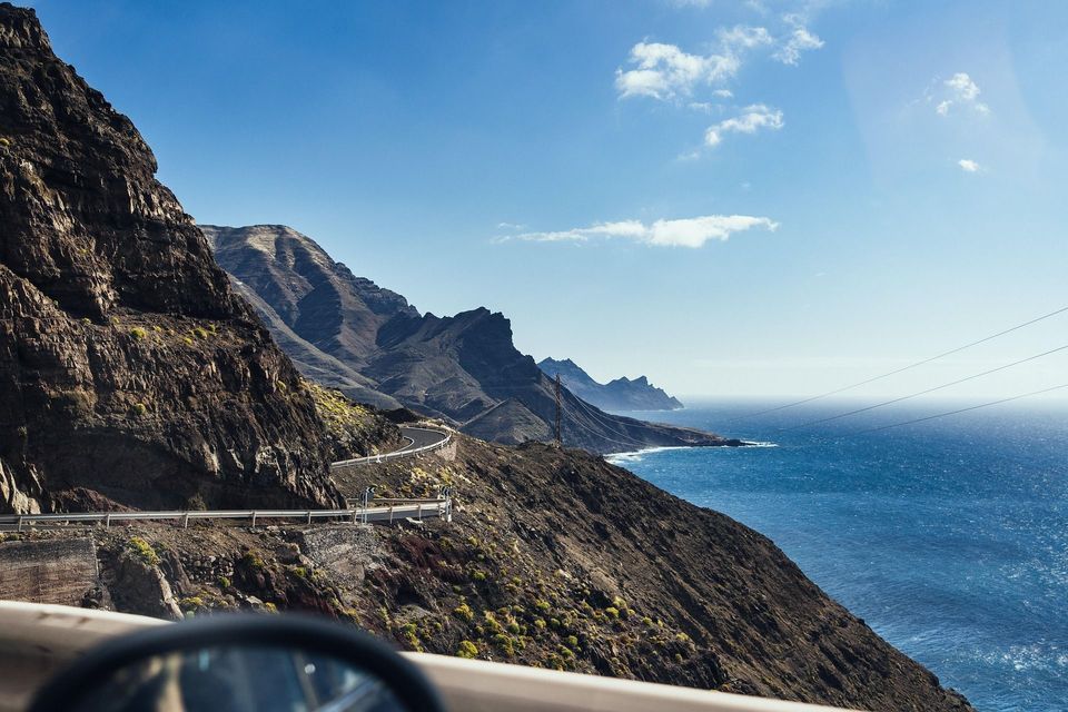 Una vista dall'auto di una strada tortuosa lungo una costa ripida e rocciosa accanto al mare blu profondo sotto un cielo soleggiato.