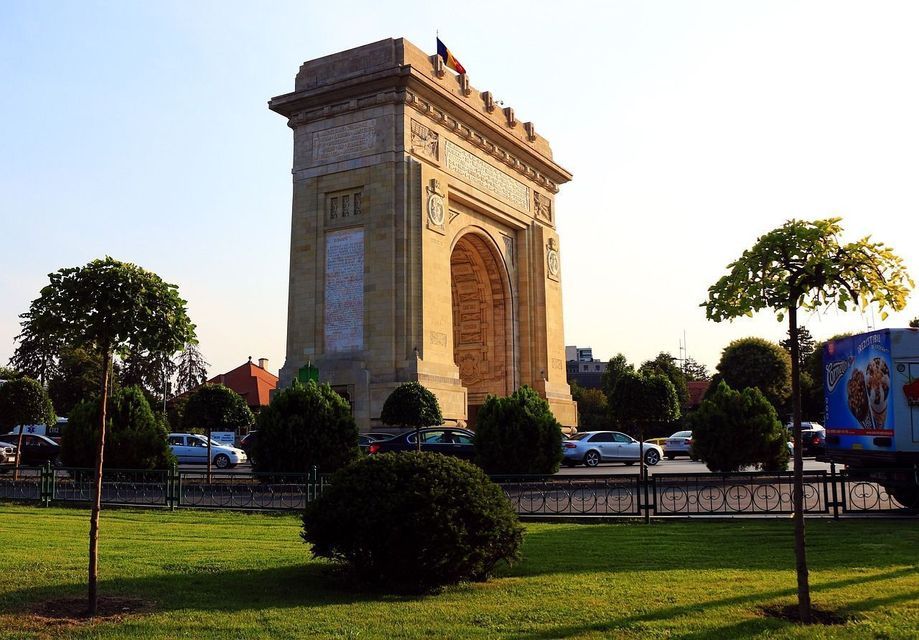 Un gran arco de triunfo de piedra se alza tras un verde césped de parque, con coches circulando por una carretera intermedia bajo un cielo despejado.