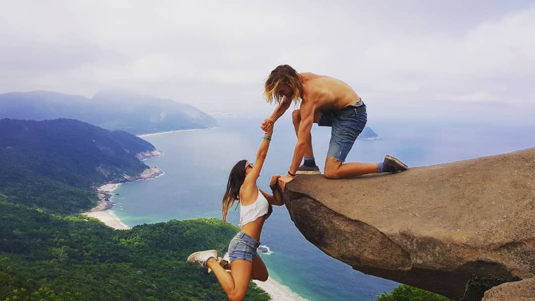 A man kneels on a rocky cliff edge, holding the hand of a woman hanging below, with a scenic coastline in the background.