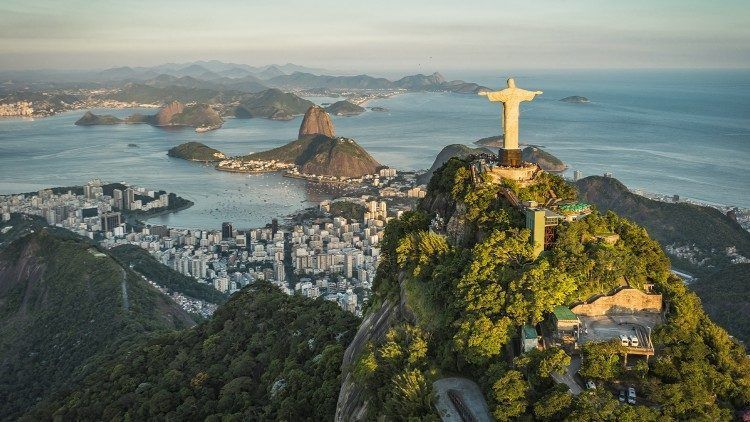 The Christ the Redeemer statue stands on a forested mountain overlooking a coastal city, a bay, and other mountains at sunset.