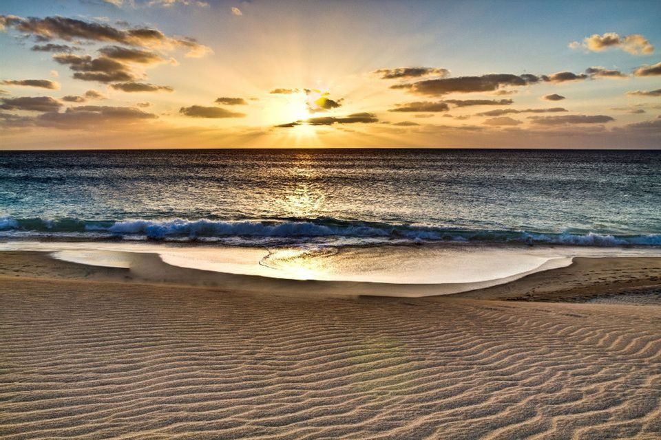 Il sole tramonta sull'orizzonte dell'oceano, i suoi raggi filtrano tra le nuvole e si riflettono sull'acqua che lambisce una spiaggia di sabbia increspata.
