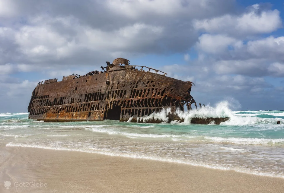 Un grande relitto arrugginito si trova arenato su una spiaggia sabbiosa mentre onde turchesi si infrangono contro il suo scafo in decomposizione sotto un cielo parzialmente nuvoloso.