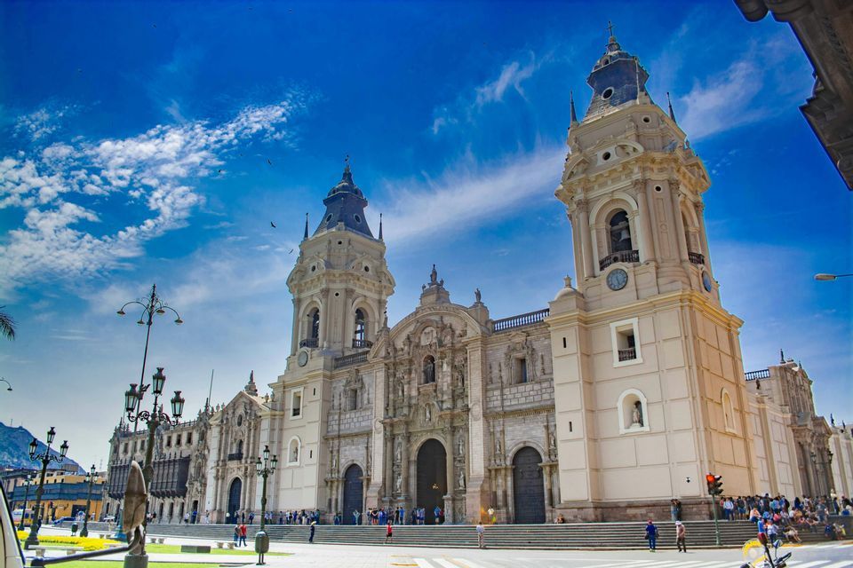 Una cattedrale ornata con due grandi campanili si erge in una piazza cittadina sotto un cielo azzurro e parzialmente nuvoloso.
