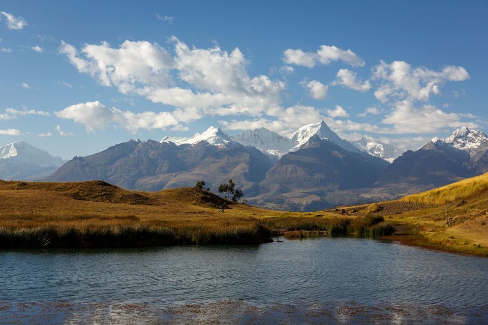 Un lago di montagna si trova in una valle erbosa di fronte a una lontana catena montuosa innevata sotto un cielo parzialmente nuvoloso.