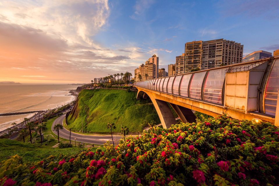 Un ponte pedonale coperto su una scogliera verde che si affaccia su una città costiera e sull'oceano durante un tramonto colorato.
