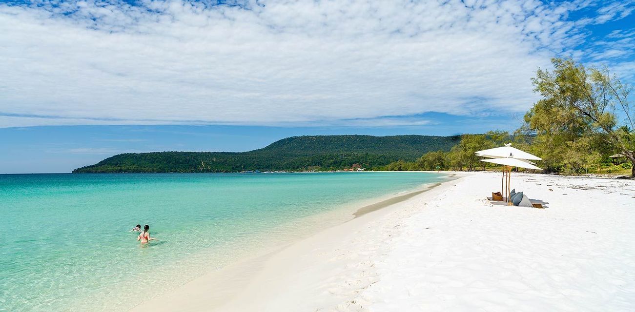 Una vasta spiaggia di sabbia bianca incontra acque calme e turchesi sotto un cielo azzurro, con due ombrelloni sulla riva e una collina boscosa in lontananza.