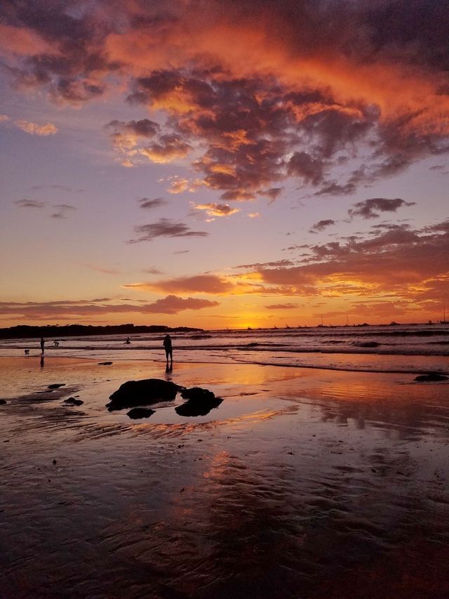 Des silhouettes de personnes sur une plage mouillée à marée basse, avec un coucher de soleil aux couleurs orange et violet qui se reflète sur le sable et l'eau.