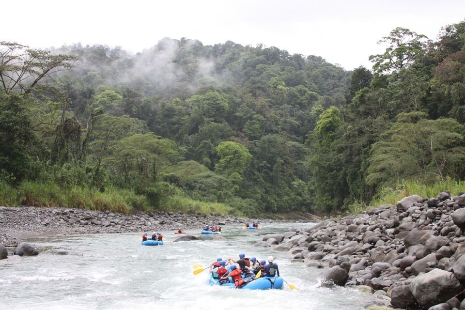 Une aventure WeRoad en groupe : du rafting en raft bleu sur une rivière bordée d'une jungle dense et brumeuse.