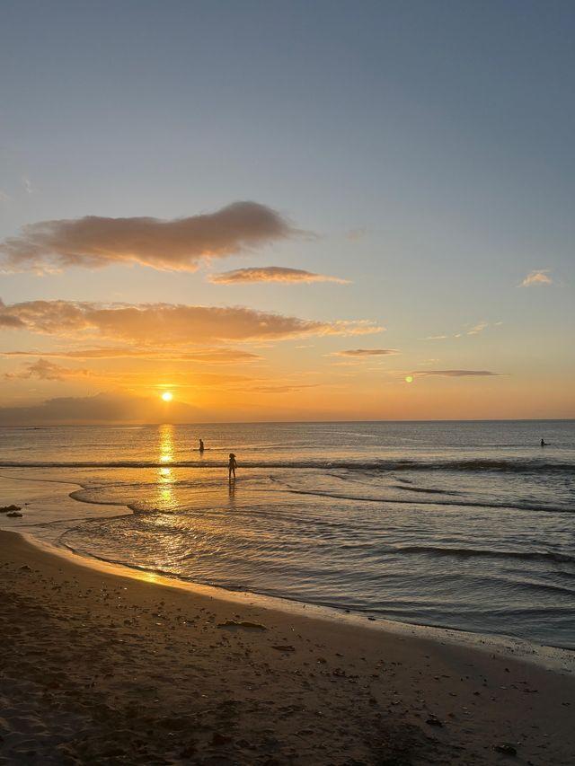 Silhouettes de trois personnes se tenant dans l'océan sur une plage de sable au coucher du soleil, projetant un reflet doré sur l'eau.