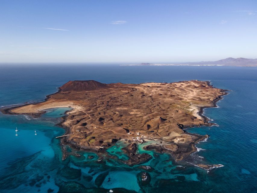 Una vista aerea di un'isola vulcanica circondata da acqua turchese, con alcune barche a vela ancorate vicino alla sua costa rocciosa.