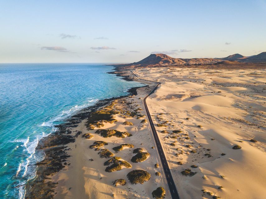 Una veduta aerea di una lunga strada che si snoda tra vaste dune di sabbia e una costa oceanica turchese, con montagne in lontananza.