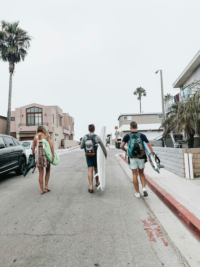 Un gruppo WeRoad di tre persone, viste di spalle, cammina per strada con le tavole da surf.