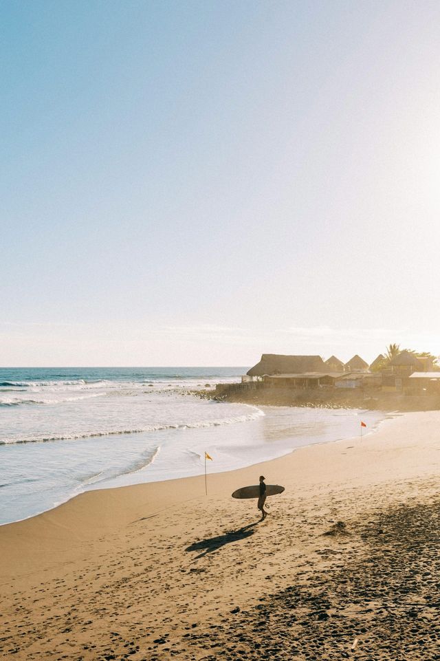 Un surfista in controluce cammina su una spiaggia sabbiosa con la sua tavola da surf, sullo sfondo l'oceano e le capanne balneari.