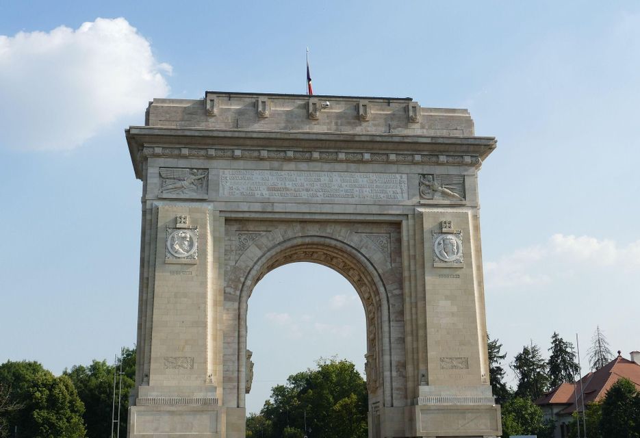 Un imponente arco trionfale in pietra, con intagli ornamentali e una bandiera in cima, si erge contro un cielo azzurro con nuvole sparse.