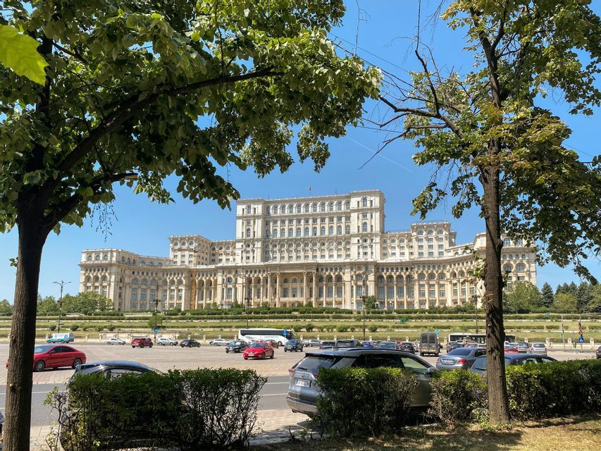 Un grande e sontuoso edificio che ricorda un palazzo, visto da oltre una strada con auto, incorniciato da alberi verdi e frondosi sotto un cielo soleggiato.