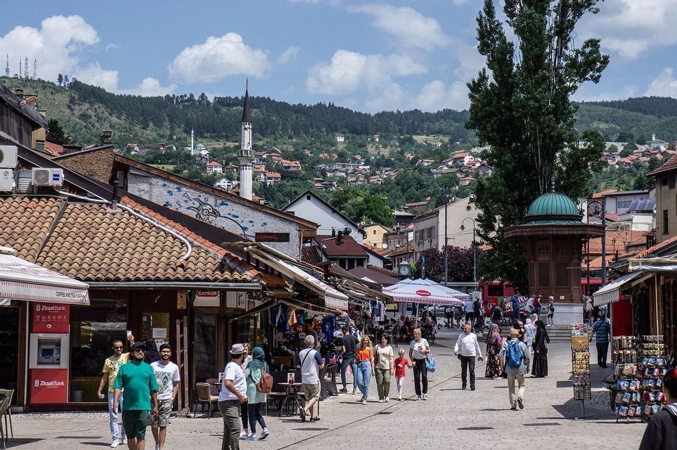 Una strada cittadina vivace e affollata di persone che passeggiano davanti ai negozi, con un minareto e colline boscose visibili sullo sfondo sotto un cielo blu.