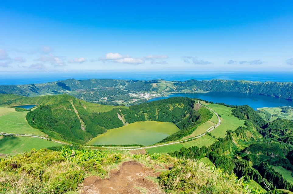 A high-angle view of green volcanic hills surrounding two crater lakes, one green and one blue, with the ocean on the horizon.