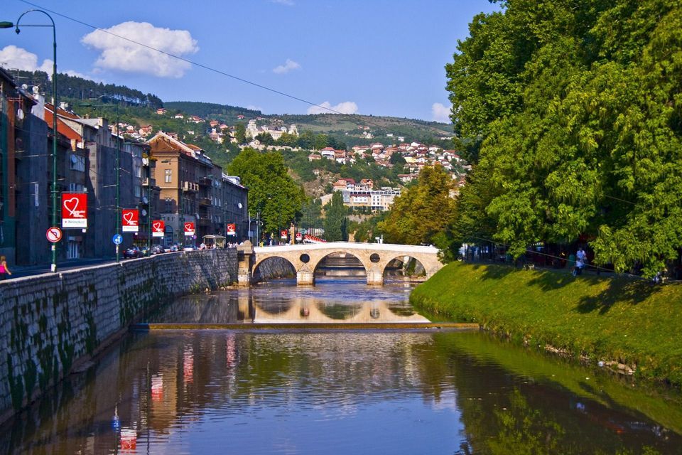 Un ponte in pietra ad arco attraversa un fiume che scorre in una città con edifici e una riva erbosa e alberata sotto un cielo azzurro e limpido.