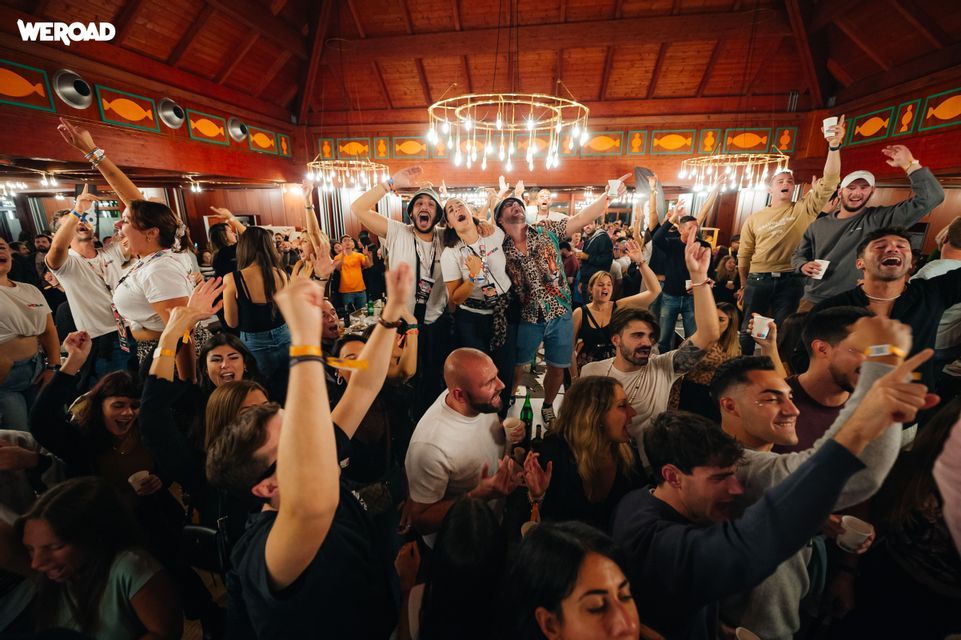 Un gruppo WeRoad esulta e balla con le mani alzate a una festa al chiuso sotto un grande lampadario.