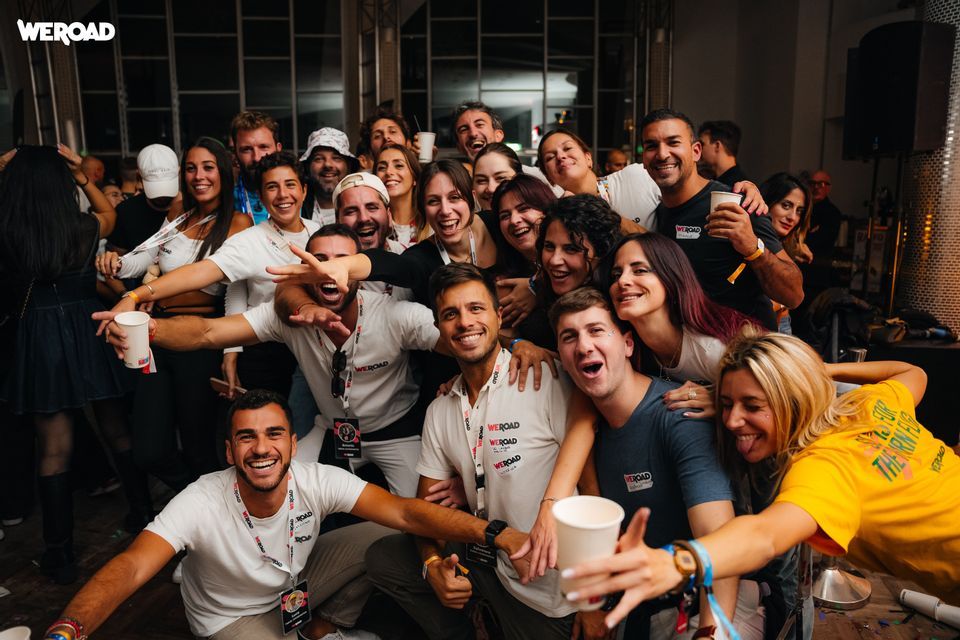 Un grande gruppo WeRoad in viaggio, con persone che sorridono e posano insieme per un selfie di gruppo a una festa al chiuso.
