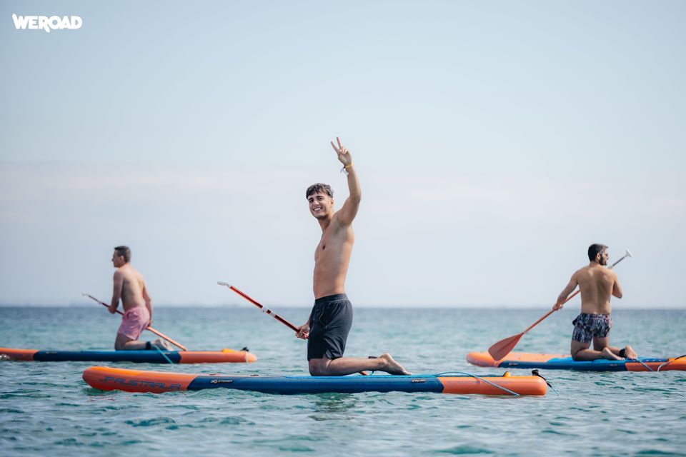 Tre uomini di un gruppo WeRoad inginocchiati su paddleboard in mare, uno sorride e fa il segno di pace.