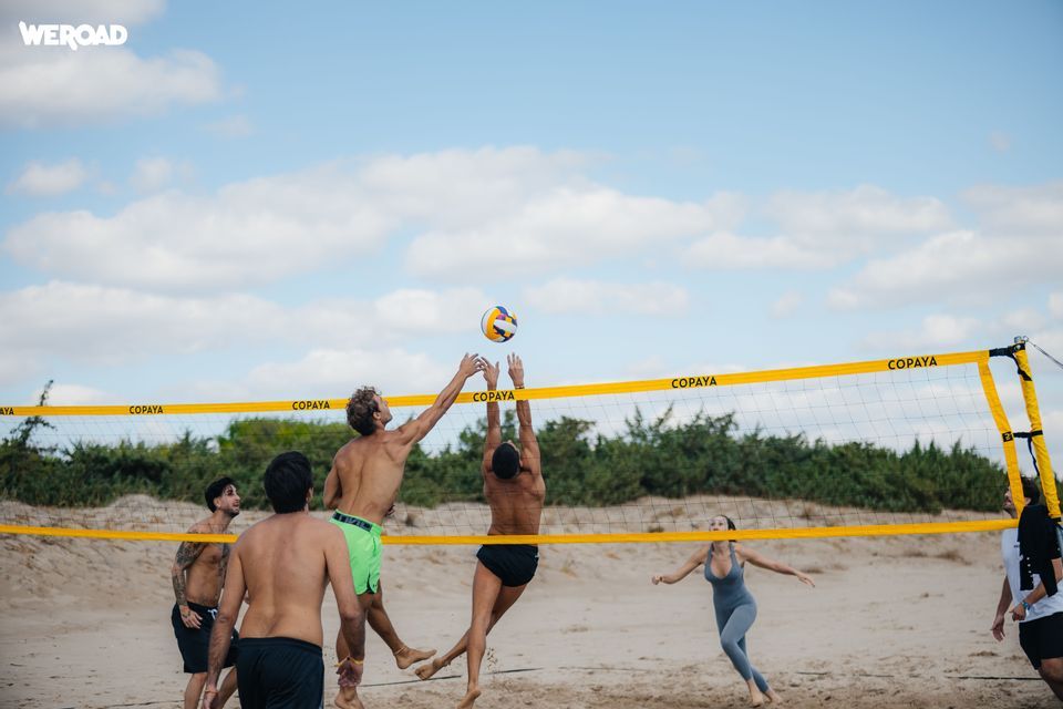 Un viaggio di gruppo WeRoad con una partita di beach volley: due uomini saltano per colpire la palla sopra la rete su una spiaggia sabbiosa.