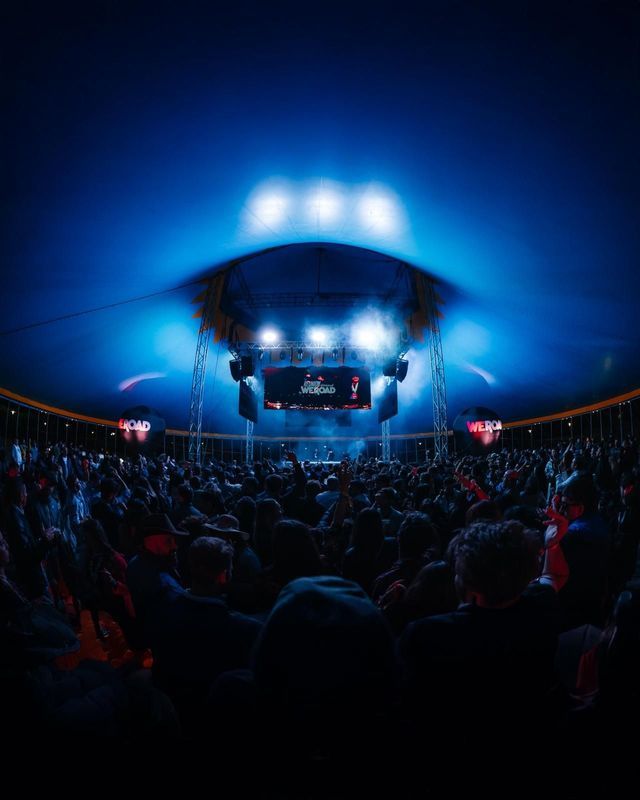 Una grande folla a un evento WeRoad, di fronte a un palco illuminato, vista da dietro sotto una vasta cupola blu.