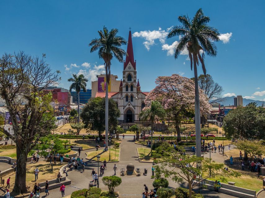 Une vue surélevée d'un parc urbain animé avec des gens, des palmiers et une église au clocher rouge en arrière-plan sous un ciel bleu.