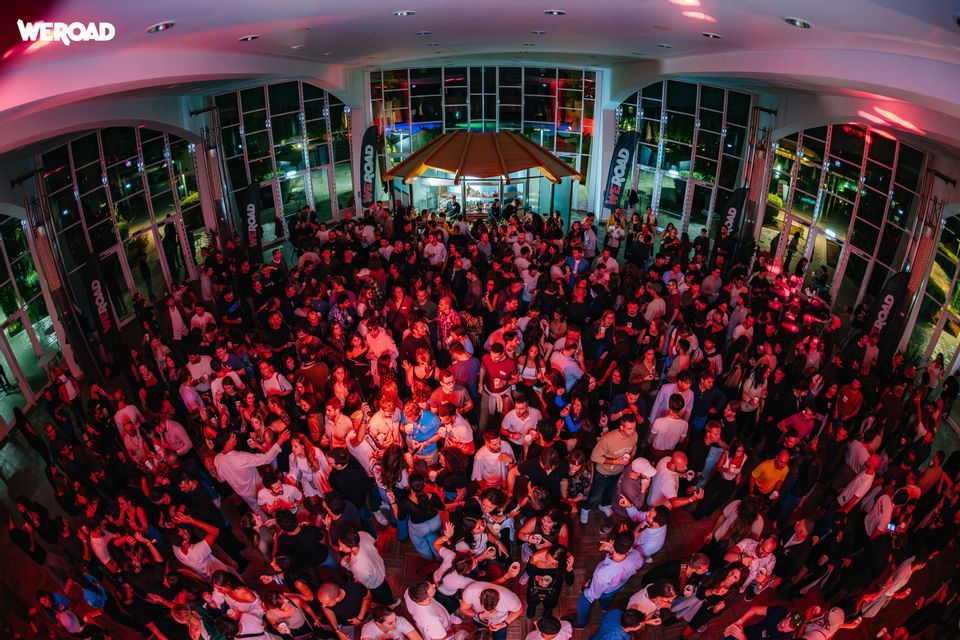 Vista dall'alto, fish-eye, di una grande folla che balla e socializza a un evento WeRoad al chiuso con illuminazione rossa.