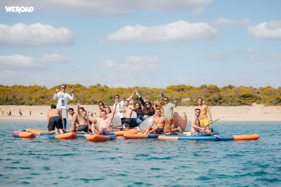 Un viaggio di gruppo WeRoad in posa insieme su paddleboard nel mare vicino a una spiaggia sabbiosa sotto un cielo parzialmente nuvoloso.