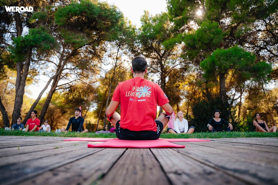 Un gruppo WeRoad in viaggio si trova in posa meditativa in un parco, visto da dietro un uomo su un tappetino da yoga.