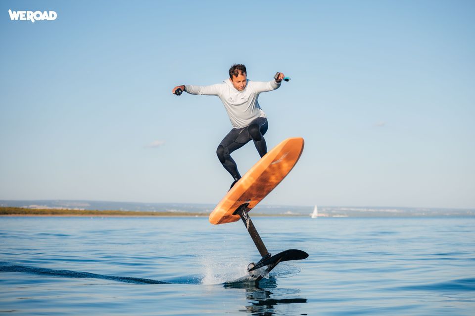 Un uomo in muta cavalca una tavola da surf idrofoil elettrica arancione, levitando sopra l'acqua calma sotto un cielo azzurro e limpido.