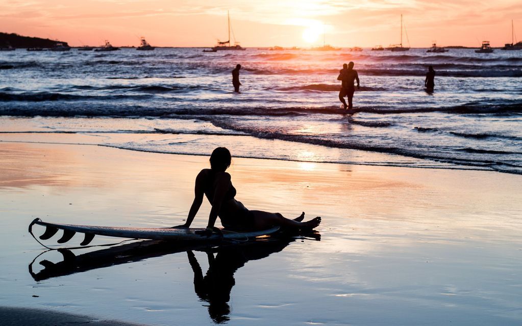 Une femme en silhouette est assise sur une planche de surf sur le sable mouillé d'une plage au coucher du soleil, avec des bateaux à l'horizon.