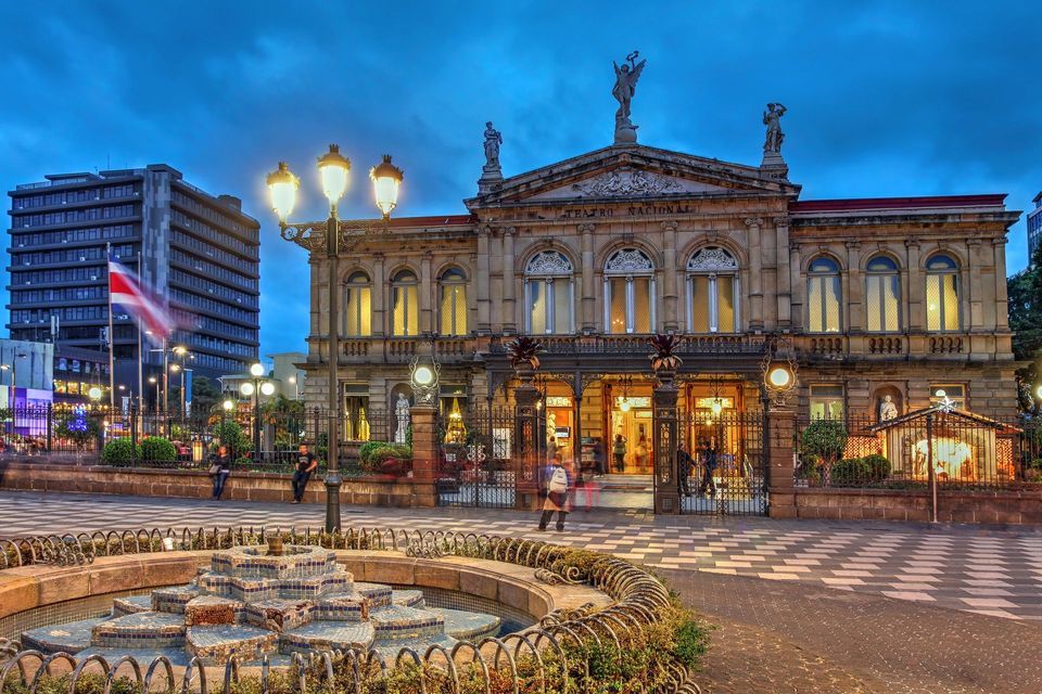 El ornamentado edificio del Teatro Nacional de Costa Rica, iluminado al atardecer, con una fuente decorativa en primer plano de una plaza de la ciudad.
