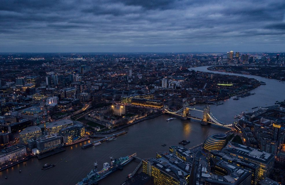 Una vista aerea del panorama urbano di Londra al tramonto, con il Tower Bridge illuminato e il Tamigi sotto un cielo nuvoloso.