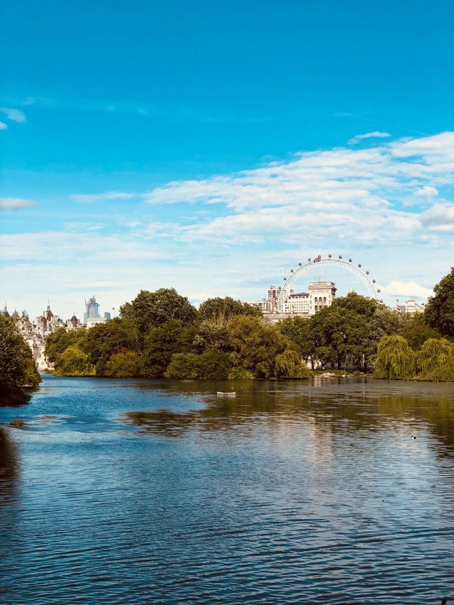 Una vista sul lago in un parco, con una grande ruota panoramica e lo skyline della città visibile dietro una fila di alberi verdi.