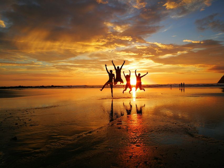 Sagome di quattro persone di un viaggio di gruppo WeRoad che saltano su una spiaggia al tramonto dorato, con riflessi sulla sabbia bagnata.