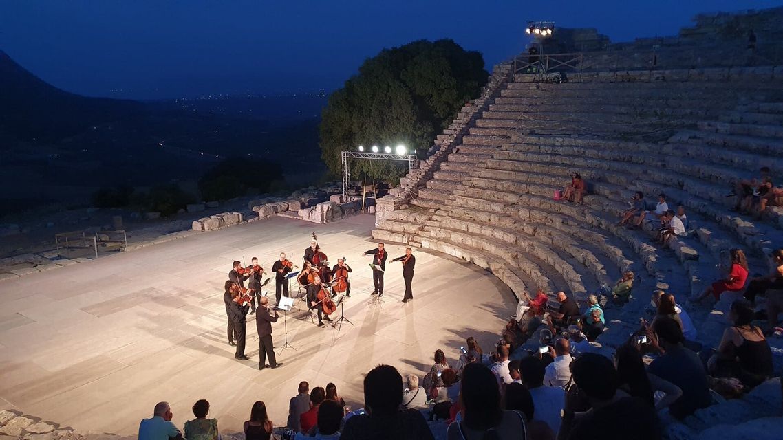 Un orchestre à cordes se produit sur scène devant un public assis dans un grand amphithéâtre de pierre au crépuscule.