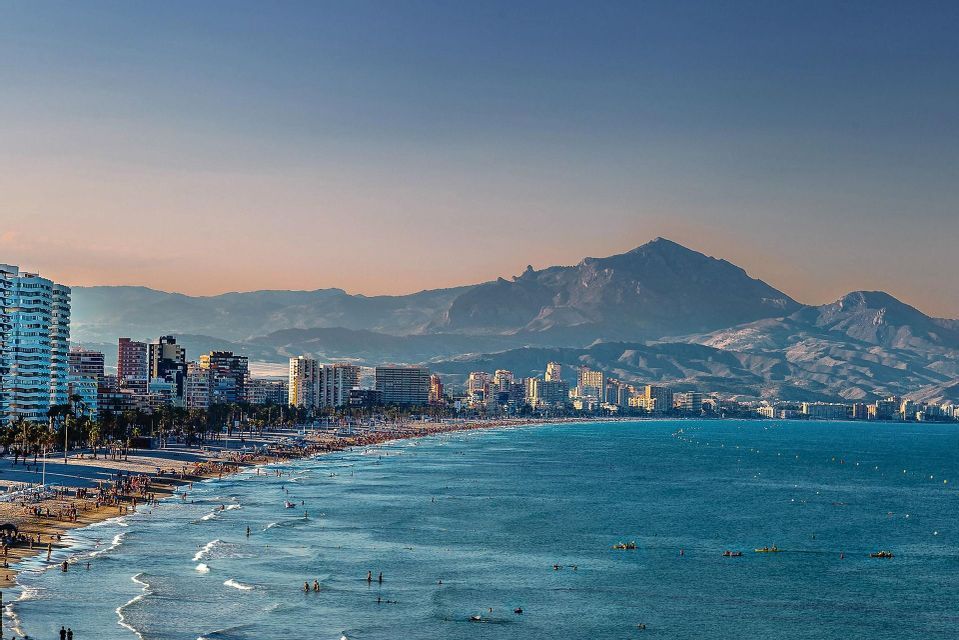 Un paesaggio urbano su una costa con una spiaggia affollata dove le persone nuotano nell'oceano, con grandi montagne visibili in lontananza.