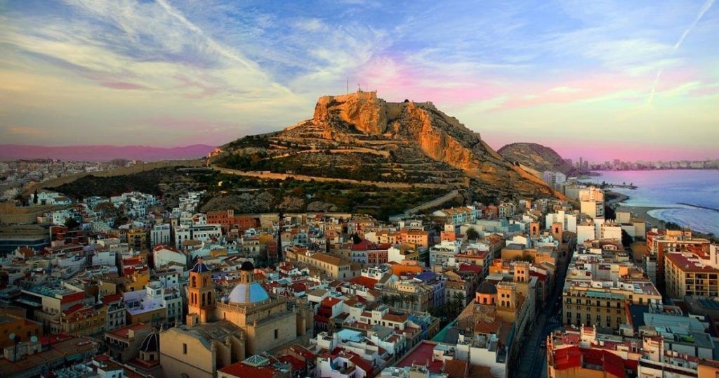 Una veduta sopraelevata di una città costiera, con un'imponente fortezza su un colle, immersa nella luce di un tramonto dai colori vivaci.